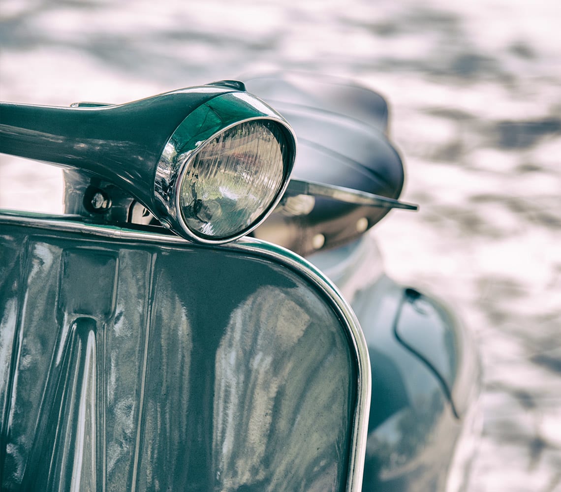 Close-up of a vintage scooter's headlight and front panel, with the seat and blurred background visible. The scooter is painted green.